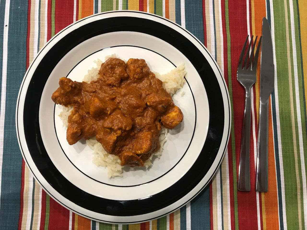 Chicken Tikka Masala over rice, plated on a white plate with black rim. Plate on rainbow tablecloth with cutlery next to it.